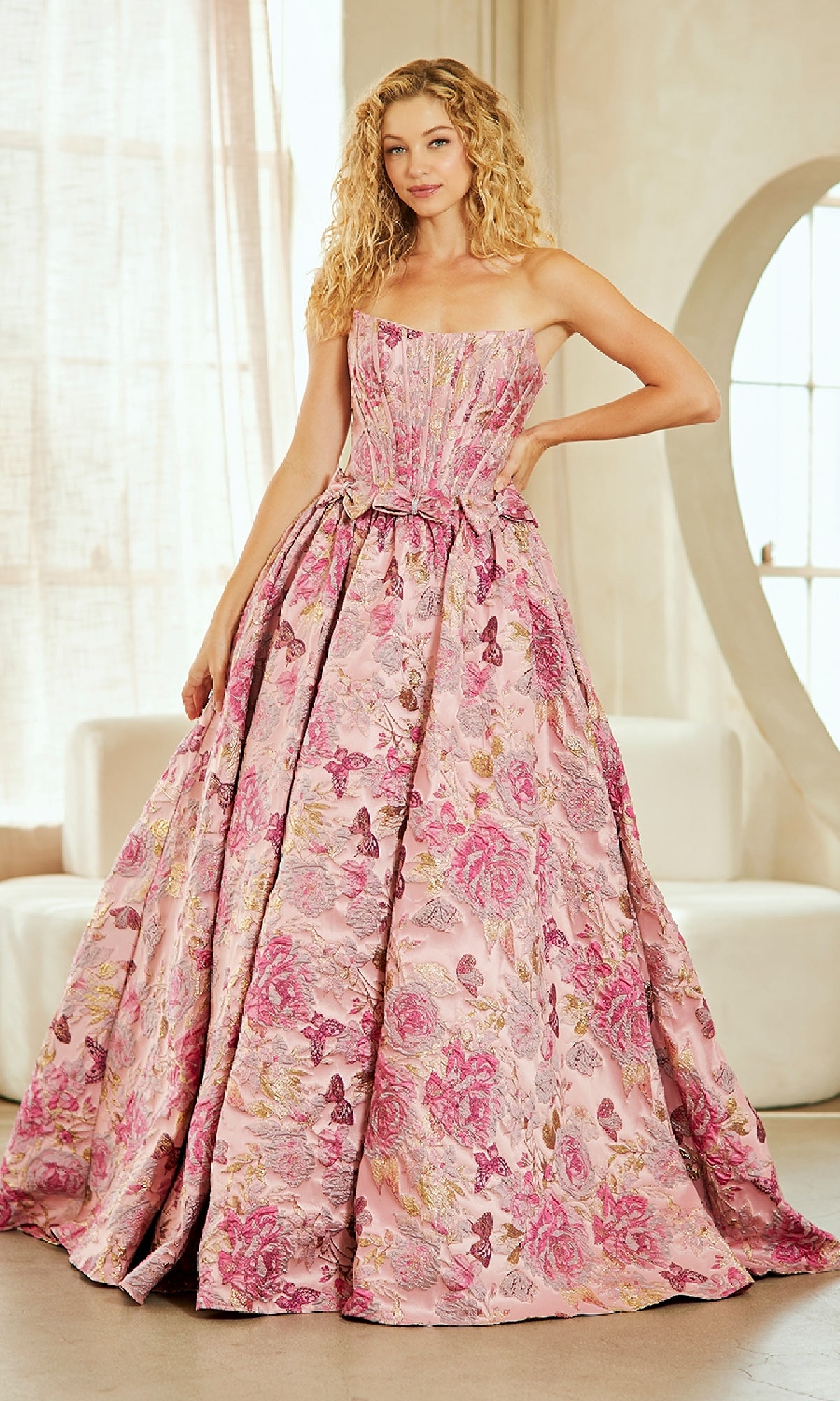 Woman standing indoors wearing the Lace-Up Print Prom Ball Gown by Amelia Couture 8068—a long pink prom dress with floral and butterfly patterns.